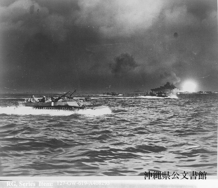 1945年。沖縄島を艦砲射撃——上陸日、海軍の大戦艦が海岸の日本軍施設めがけて集中砲火を浴びせる中、海岸警護艇に警護された揚陸艦を離れて上陸第一波、第二波と共に海岸に押し寄せる水陸両用車両。沖縄県公文書館 蔵
